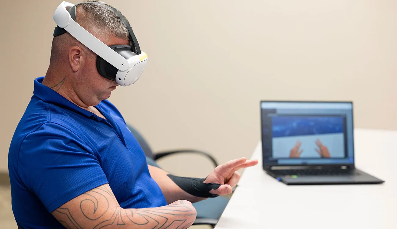 A man wearing a VR headset sits at a table during a therapy session, with a laptop showing the virtual scene.